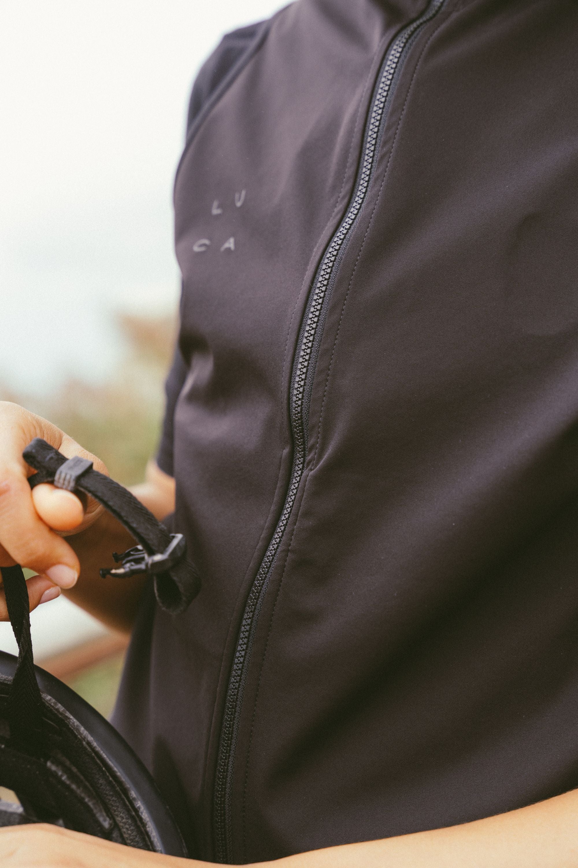 Close-up-detail-of-the-matte-black-windproof-fabric-and-full-length-black-zipper-on-the-Luca-cycling-gilet-The-subtle-dark-LUCA-logo-is-visible-on-the-chest-A-hand-holds-a-helmet-strap-in-the-foreground