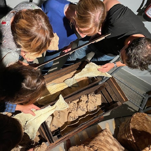 A group of people examining items over a wooden crate filled with burlap sacks, viewed from above.