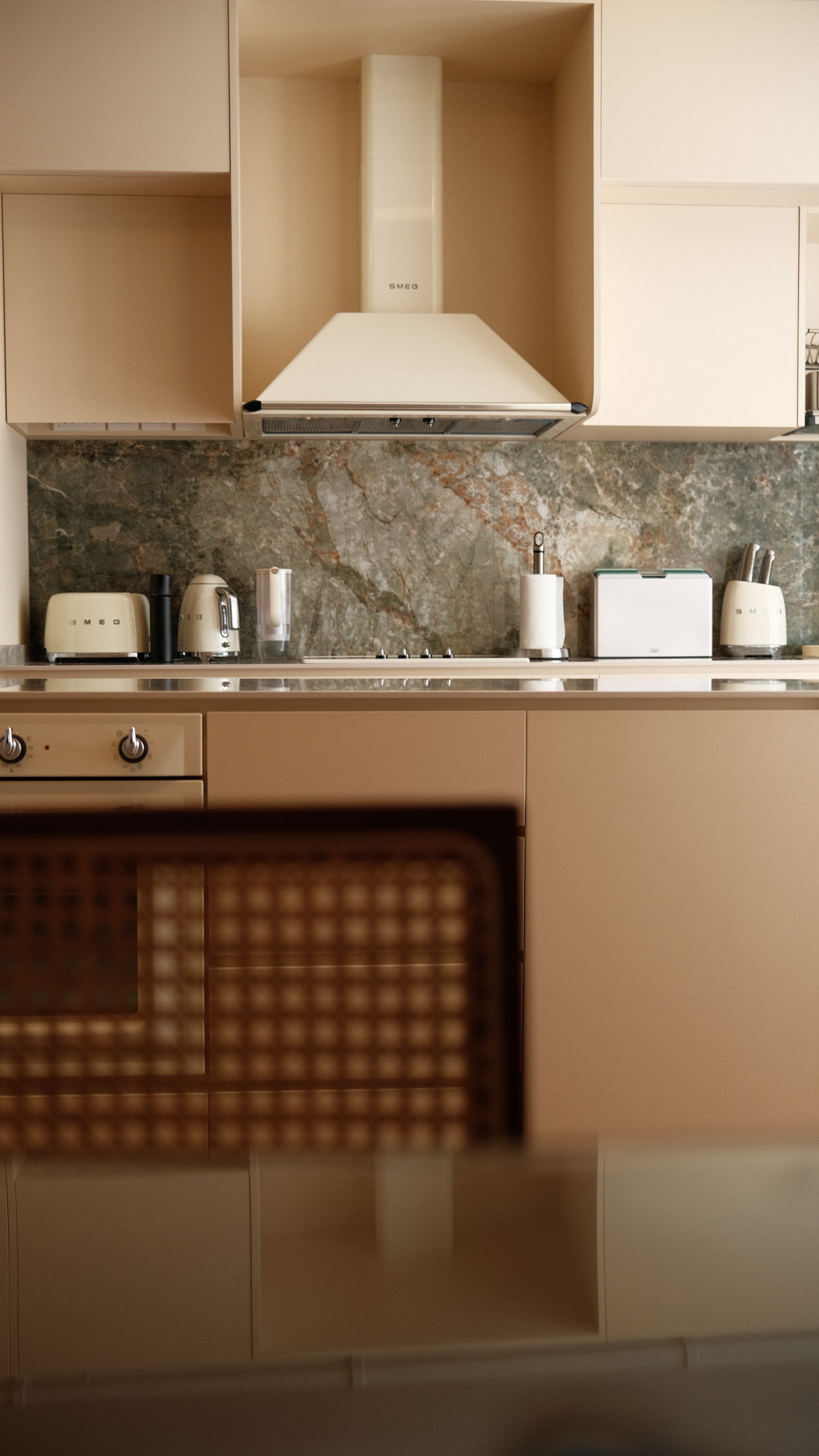 Close-up of contemporary kitchen design featuring cream-colored cabinetry, Smeg range hood, and natural stone backsplash for a timeless, elegant look.