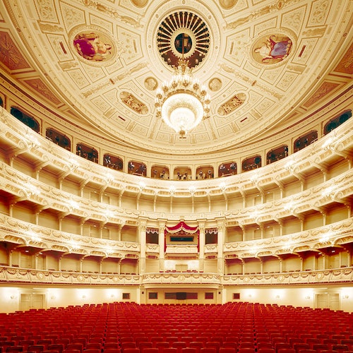 Ylellinen teatterin sisustus suurella kattokruunulla, yksityiskohtainen katto, parvekkeet ja rivissä punaisia istuimia, jotka ovat suunnattu lavalle.