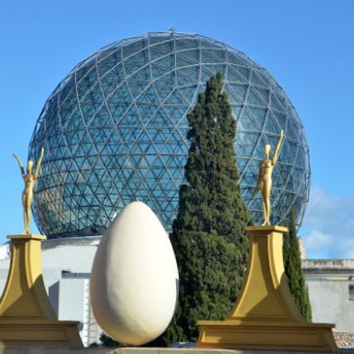 כיפה גאודזית מאחורי פסלים שמחזיקים ביצים ופסל ביצה גדול, עם עצים גבוהים ושמיים כחולים ברקע.