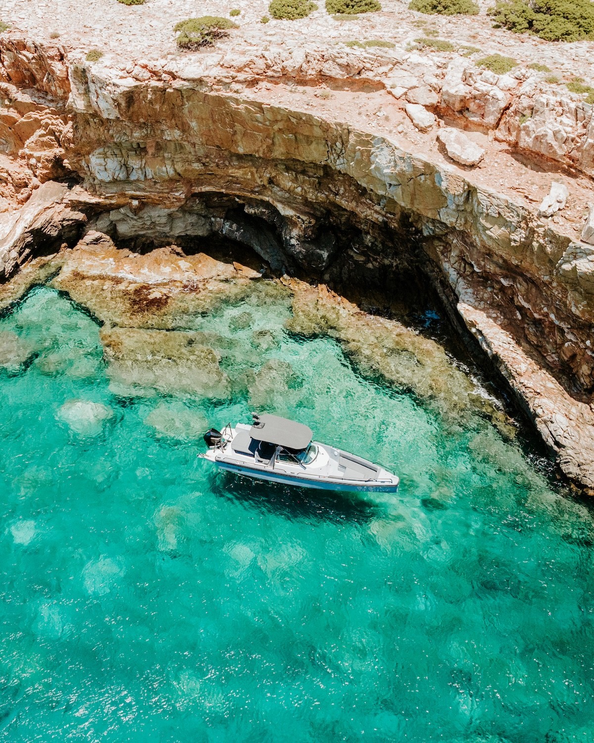 White Axopar 37 yacht anchored in crystal-clear turquoise waters near dramatic limestone cliffs and scattered rocks in a secluded Greek cove.