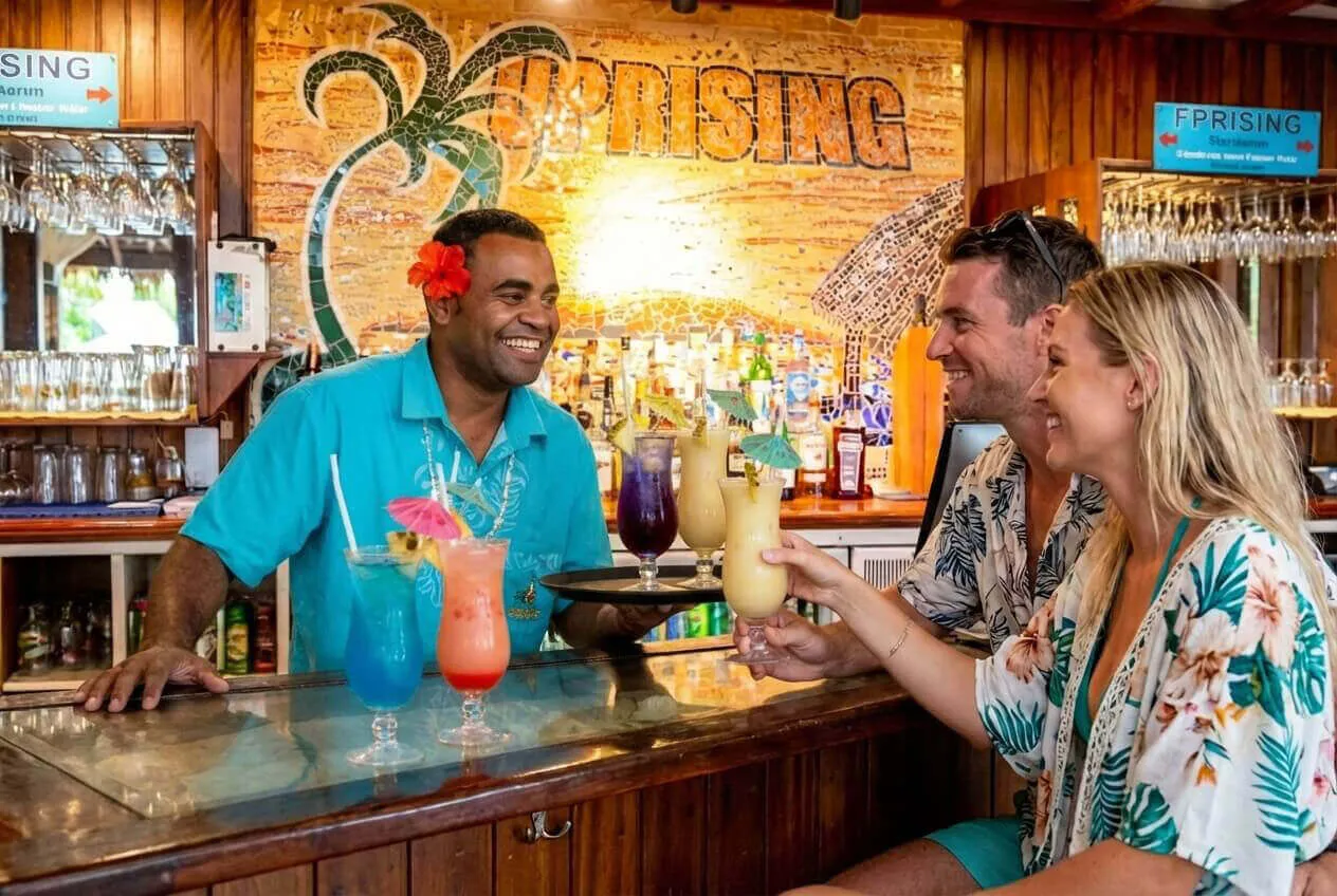 A smiling Fijian bartender serves colourful tropical drinks to a happy couple at the Uprising Beach Resort bar.