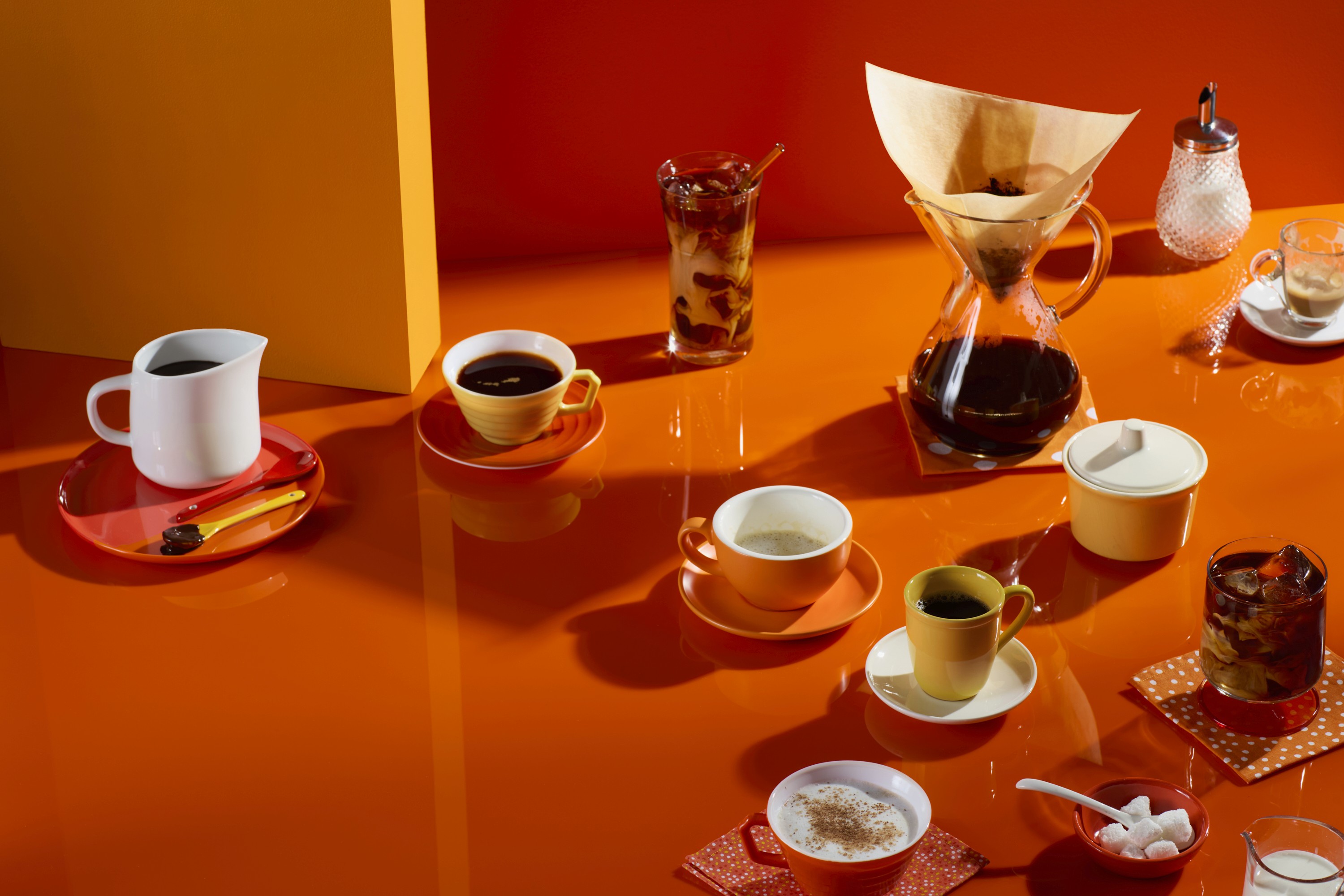 Pour-over coffee carafe with various kinds of coffee including iced coffee, latte, black coffee, cappuccino, and espresso in mismatching mugs and glasses arranged graphically on an orange background.
