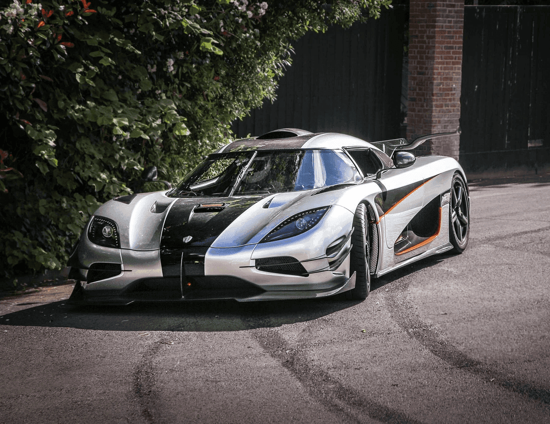 A sleek white and black sports car with an aerodynamic design is parked on a paved driveway surrounded by lush greenery, embodying luxury and speed.