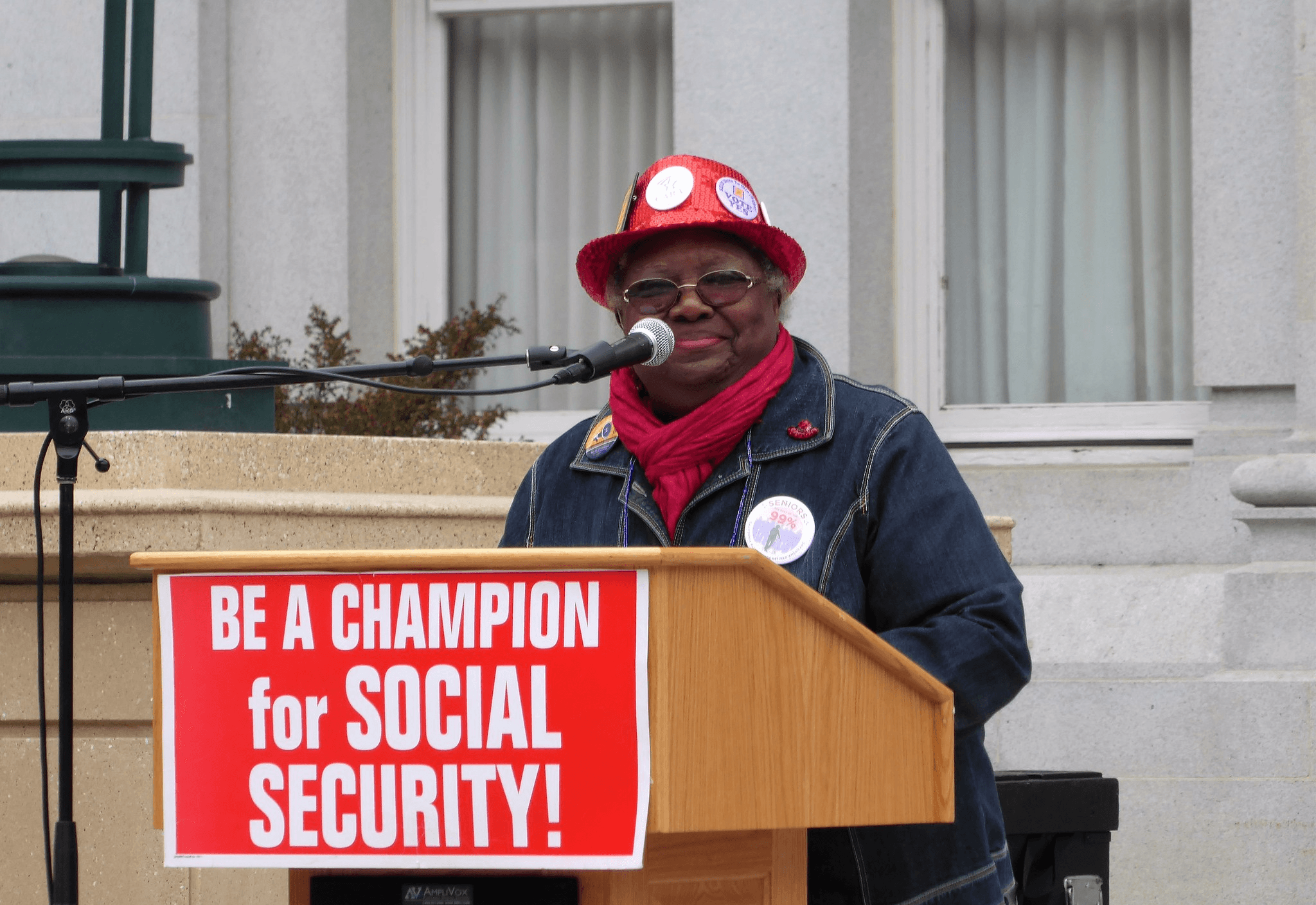 An older woman wearing a red hat and buttons speaks at a podium with a sign that reads “BE A CHAMPION for SOCIAL SECURITY!” during a public rally.