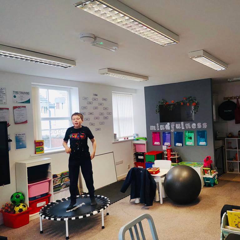 A child plays on a trampoline in a classroom, there are multiple fitness equipment items in the room for them to use.