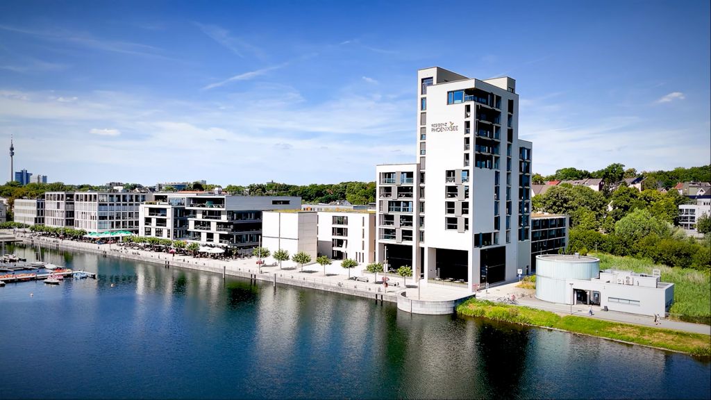 Modern residential building on waterfront with blue water, blue sky, white buildings, and landscaping visible
