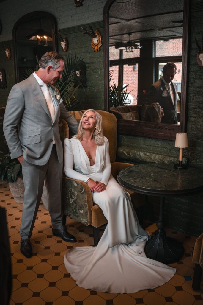 Bride seated beside her standing partner in a lounge with a dark green brick wall and large mirror