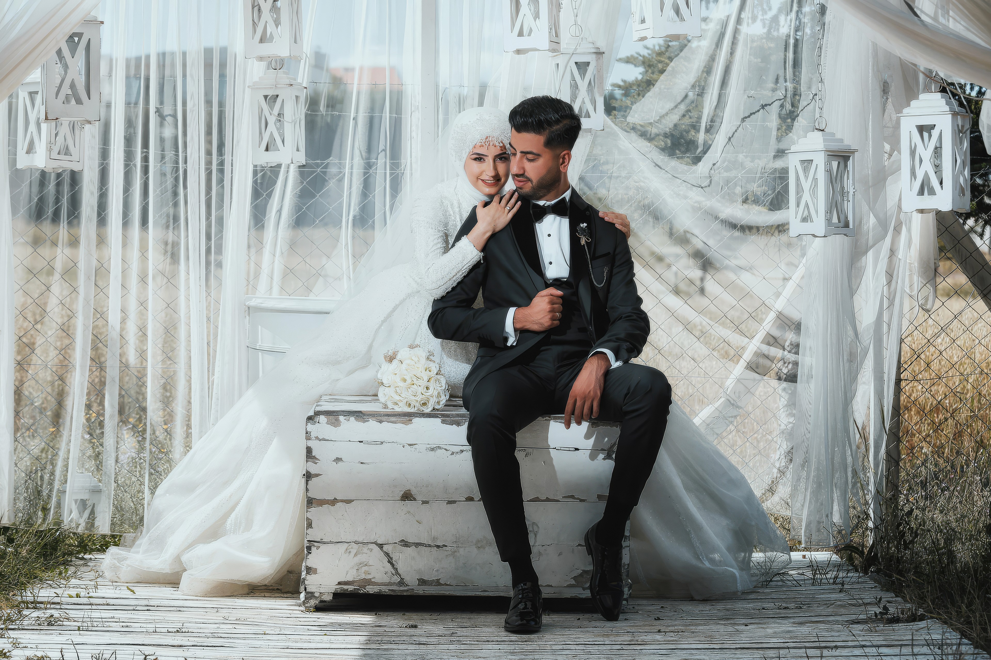 Bride in a hijab and groom in a tuxedo sitting together outdoors