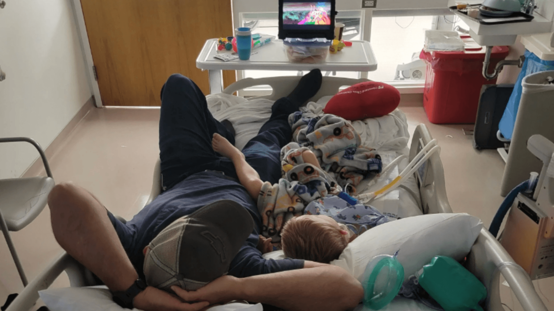 father and young son lay in hospital bed together in pediatric intensive care unit watching a movie on a tablet
