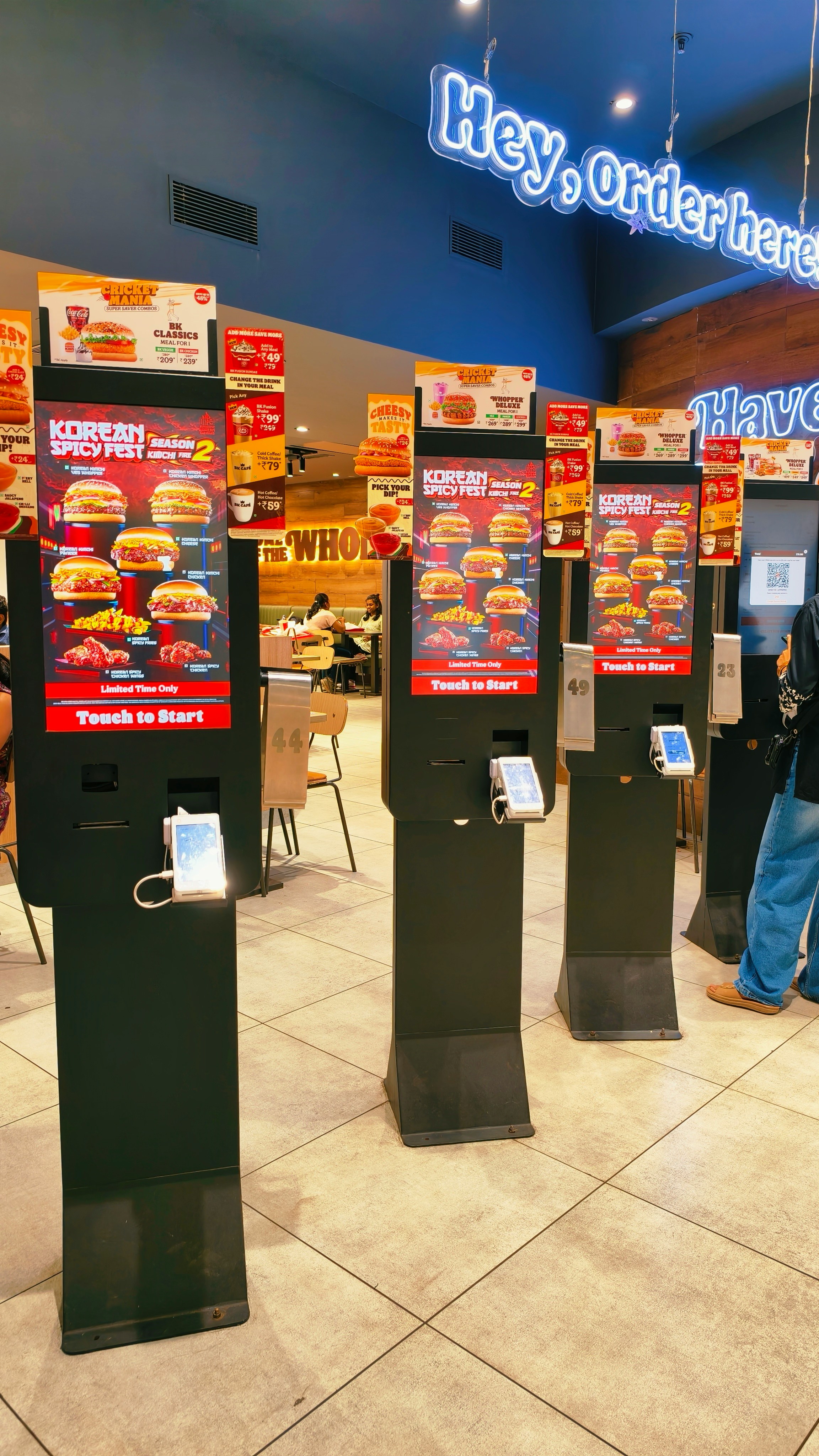 Three digital ordering kiosks in a fast food restaurant with burger menu displays