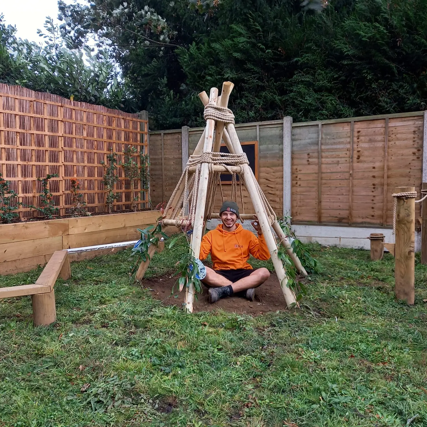 A child sits inside a wooden swing set frame in a yard, surrounded by grass and a wooden fence in the background.