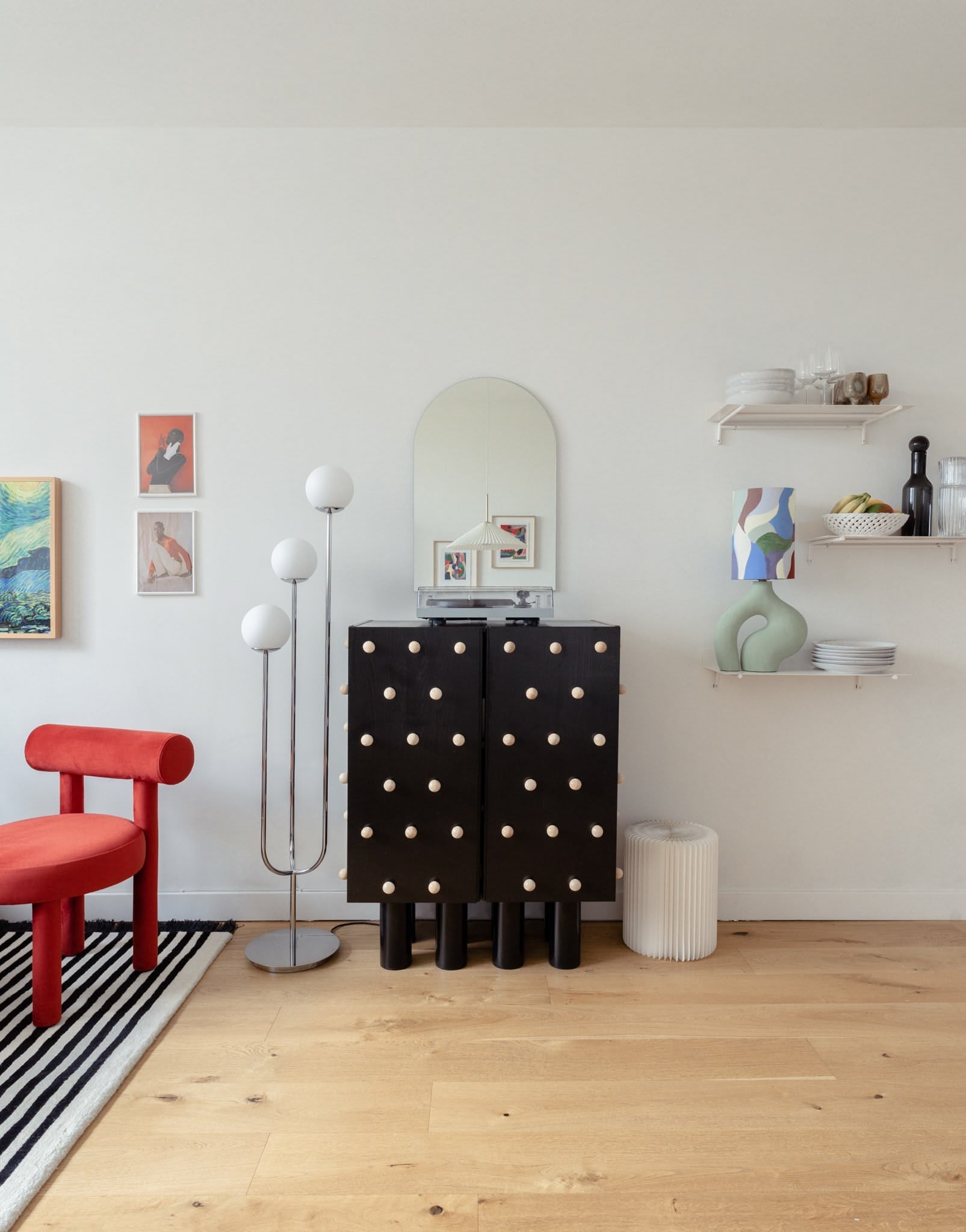 Living room with a brown detailed cabinet, decorative modern art deco lamp, framed artwork, a red armchair, and wall shelves with dishes and decorative items.