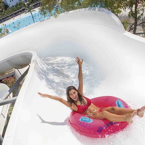 Mujer en bikini rojo sobre un flotador rosa deslizándose por un tobogán de agua blanca, con una piscina y árboles de fondo.