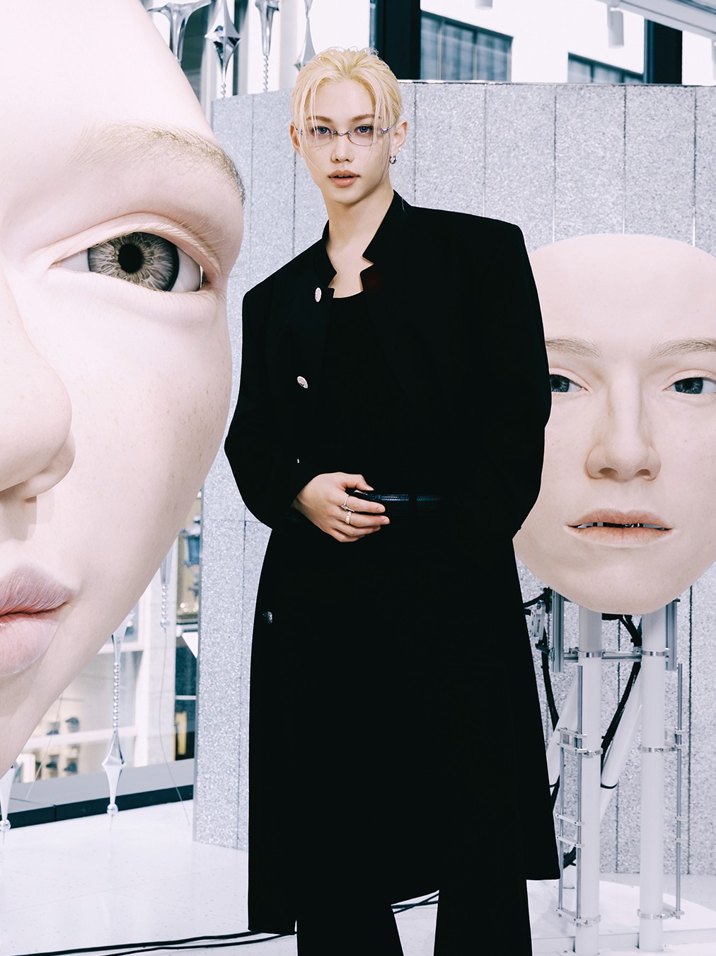 Felix of K-pop group Stray Kids wearing Gentle Monster glasses and a long black coat stands between oversized sculptural human faces in a modern, gallery-like store setting.