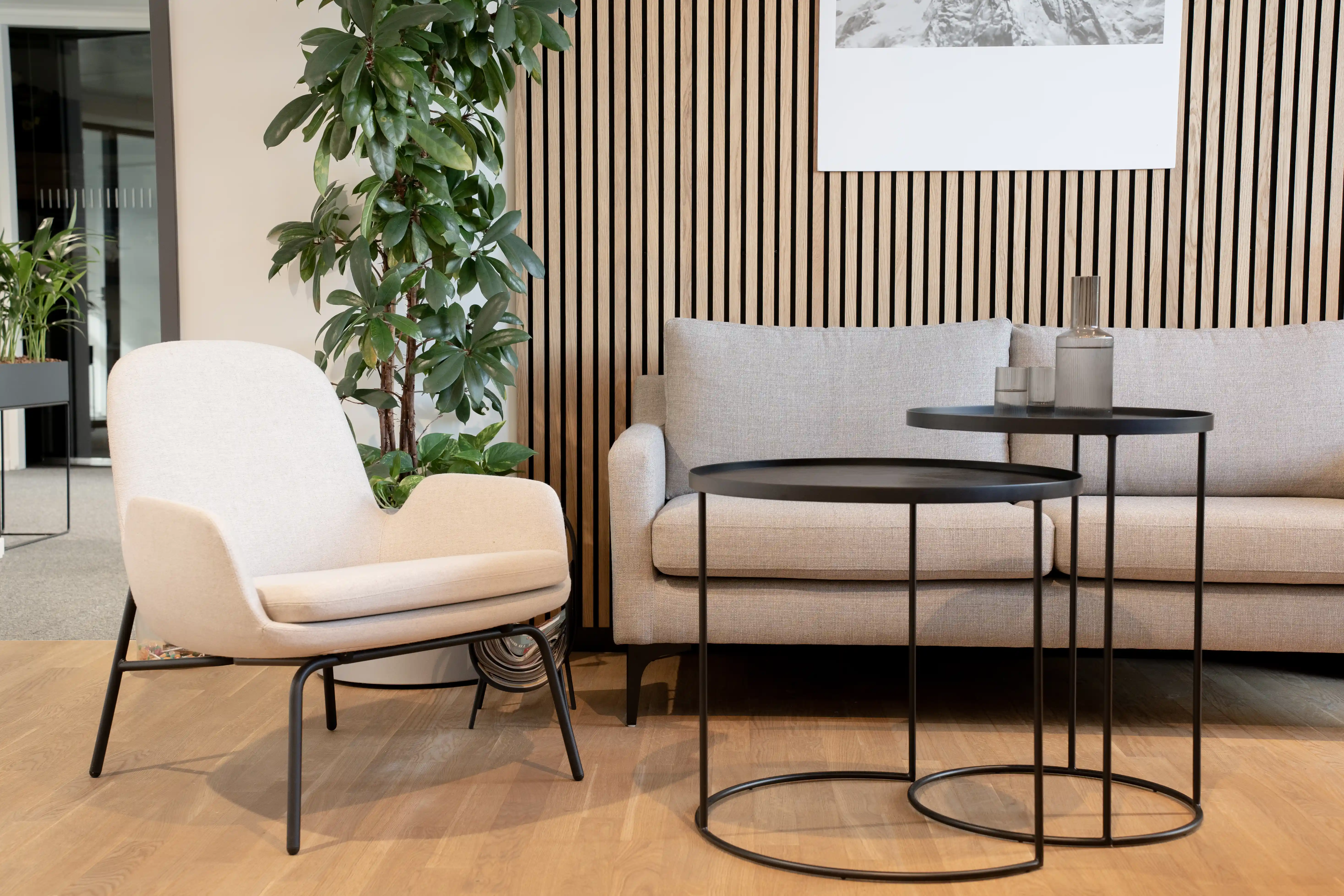 Lounge at AI START-UP MUNICH with a beige armchair and grey sofa in front of an oak acoustic slat wall, black round nesting tables with candles and a framed monochrome artwork.