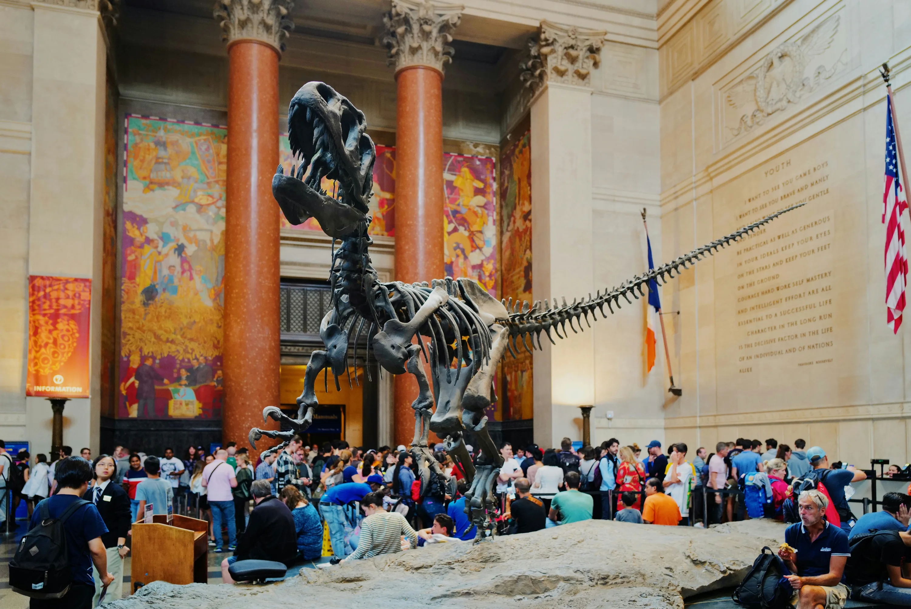 Museum visitors gaze at a towering Tyrannosaurus Rex skeleton exhibit with large columns and murals in the background.