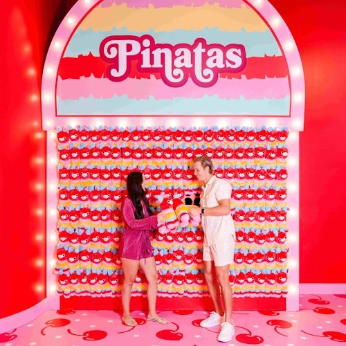 Due persone stanno di fronte a un muro pieno di pinatas a tema ciliegia. Il muro è illuminato e etichettato "Pinatas." Stanno tenendo delle pinatas.