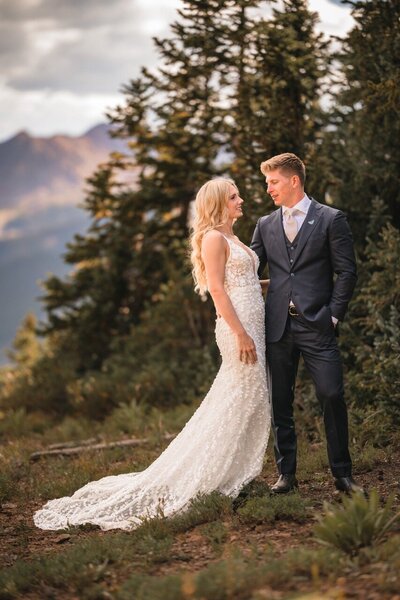 The couple stand close together among evergreens, with the bride's lace train on the ground.