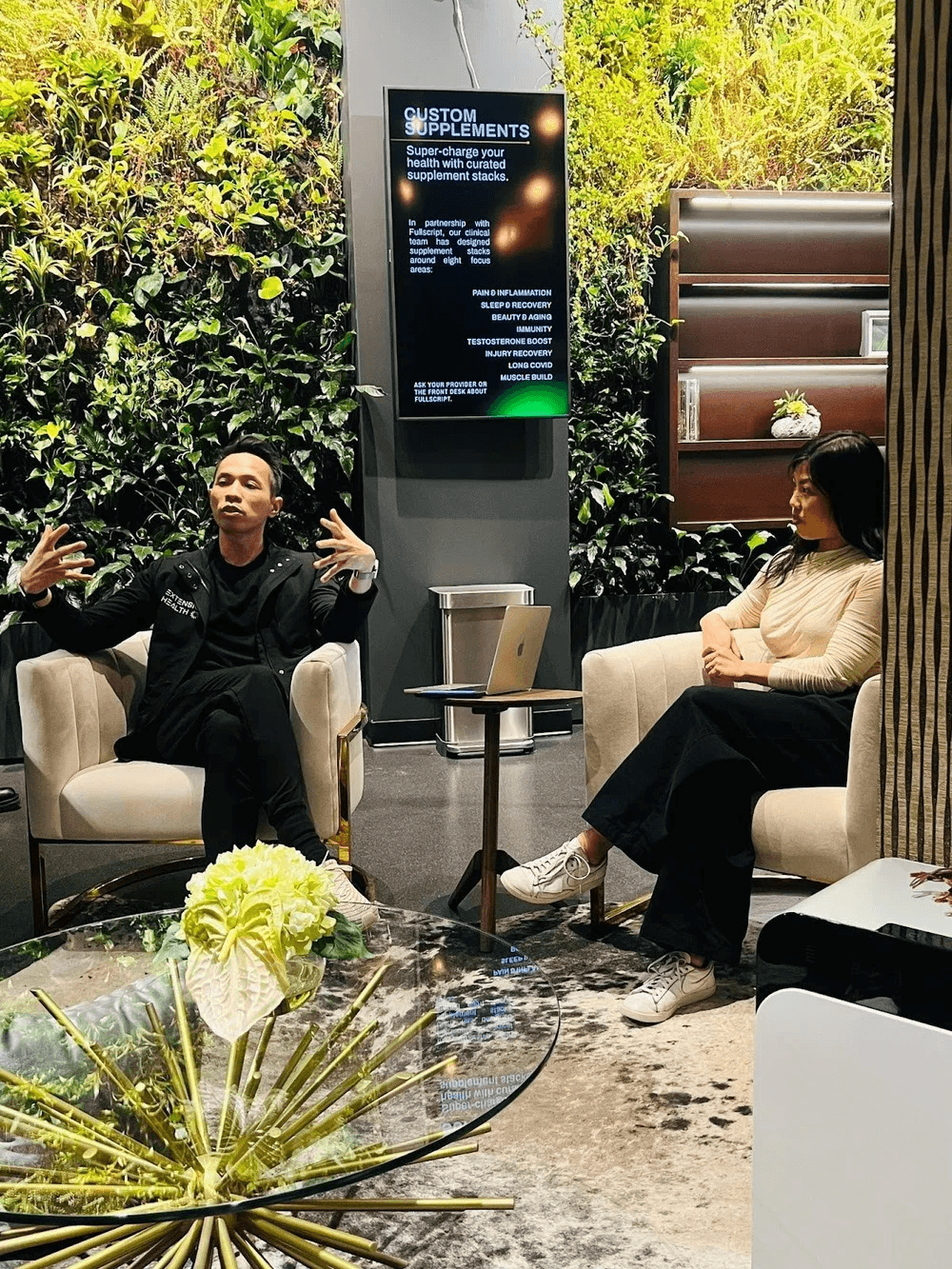 Two people seated in a lush outdoor setting, engaged in conversation next to a digital display.