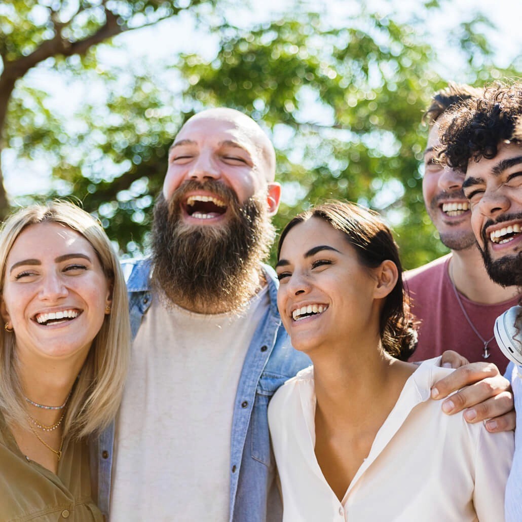 A group of five friends stand closely together outdoors,  laughing joyfully