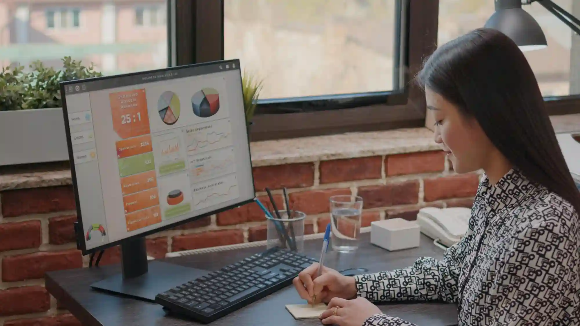 Asian businesswoman reviewing pie charts and sales reports on a desktop computer, taking notes for a business strategy.