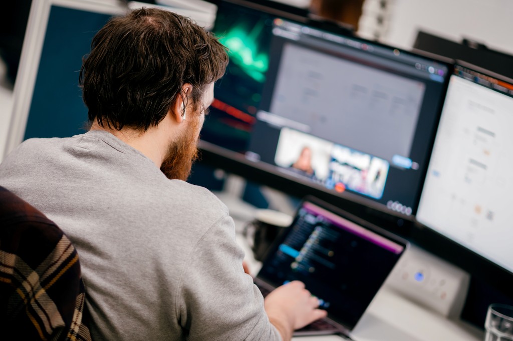 A bearded person working on a laptop, using multiple monitors with code and a video call in view.