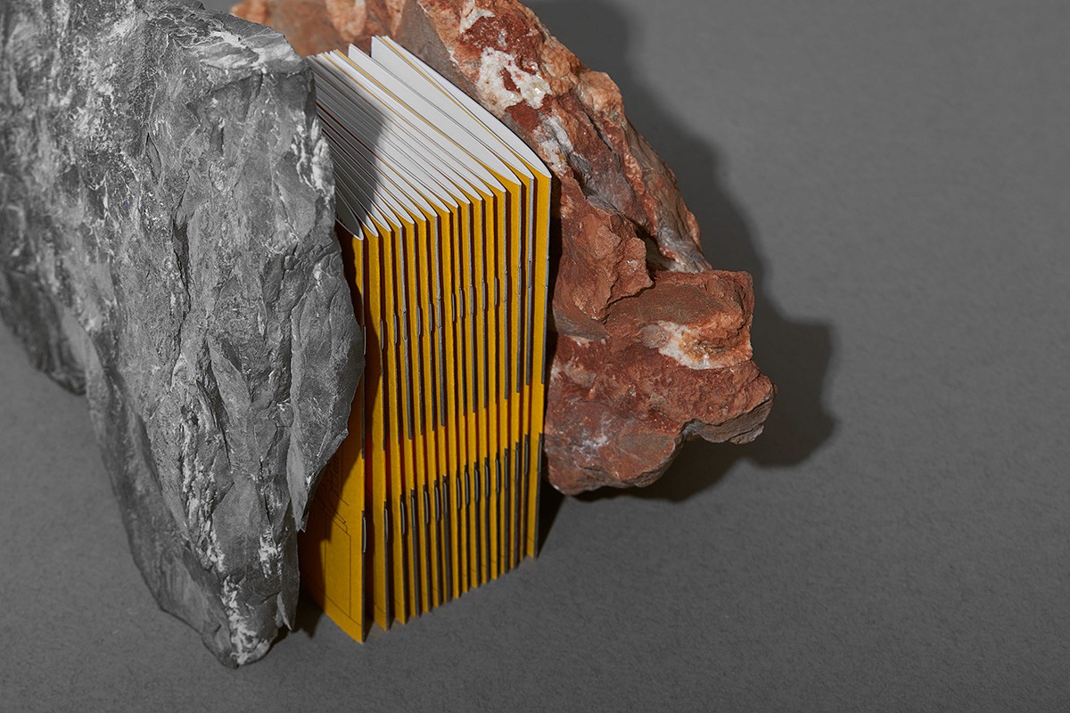 A side-angle perspective of a stack of vibrant yellow exhibition catalogues for the August Zamoyski showcase, vertically wedged between two large, textured rock fragments. This visual highlights the studio's expertise in editorial design and tactile brand experiences, using raw materials to reinforce the sculptural theme of the cultural exhibition.
