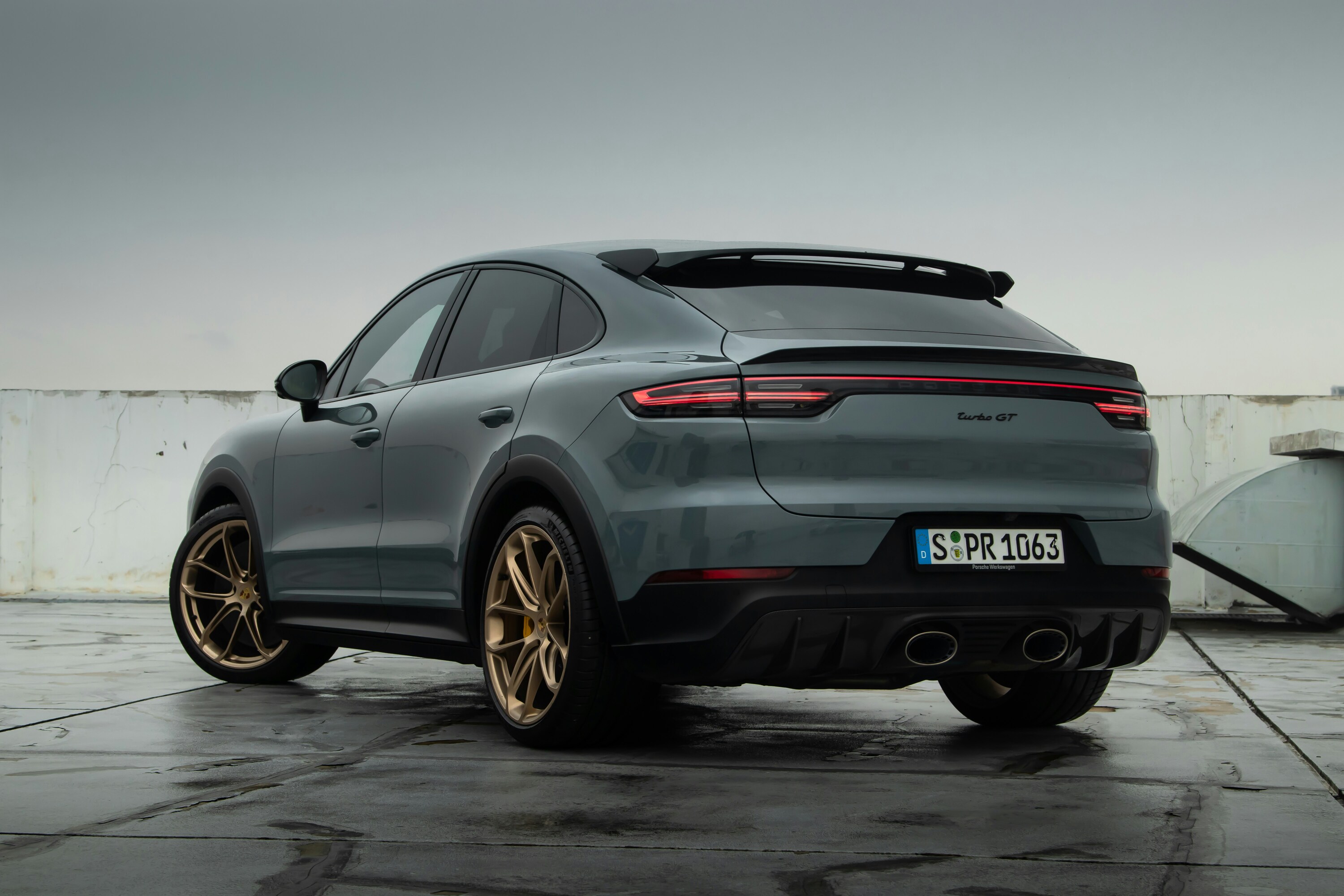 A sleek, gray luxury SUV with bronze alloy wheels is parked on a wet concrete surface under a cloudy sky, showcasing its modern design and aerodynamic contours.