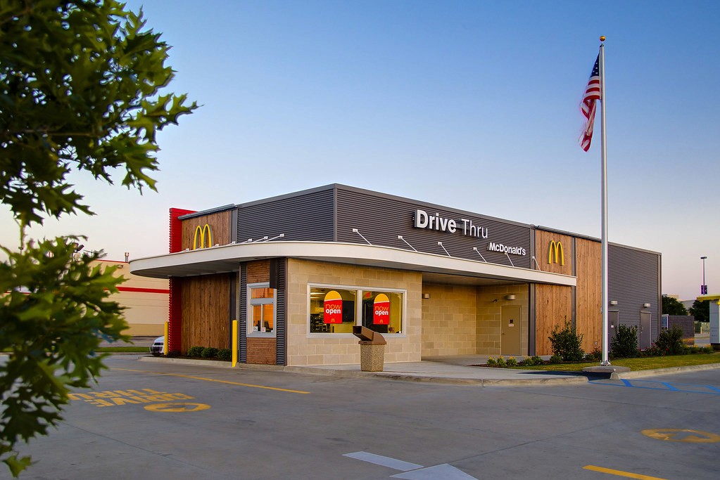 A modern building with a flat roof, trees in front, and an American flag on a pole against a clear sky.