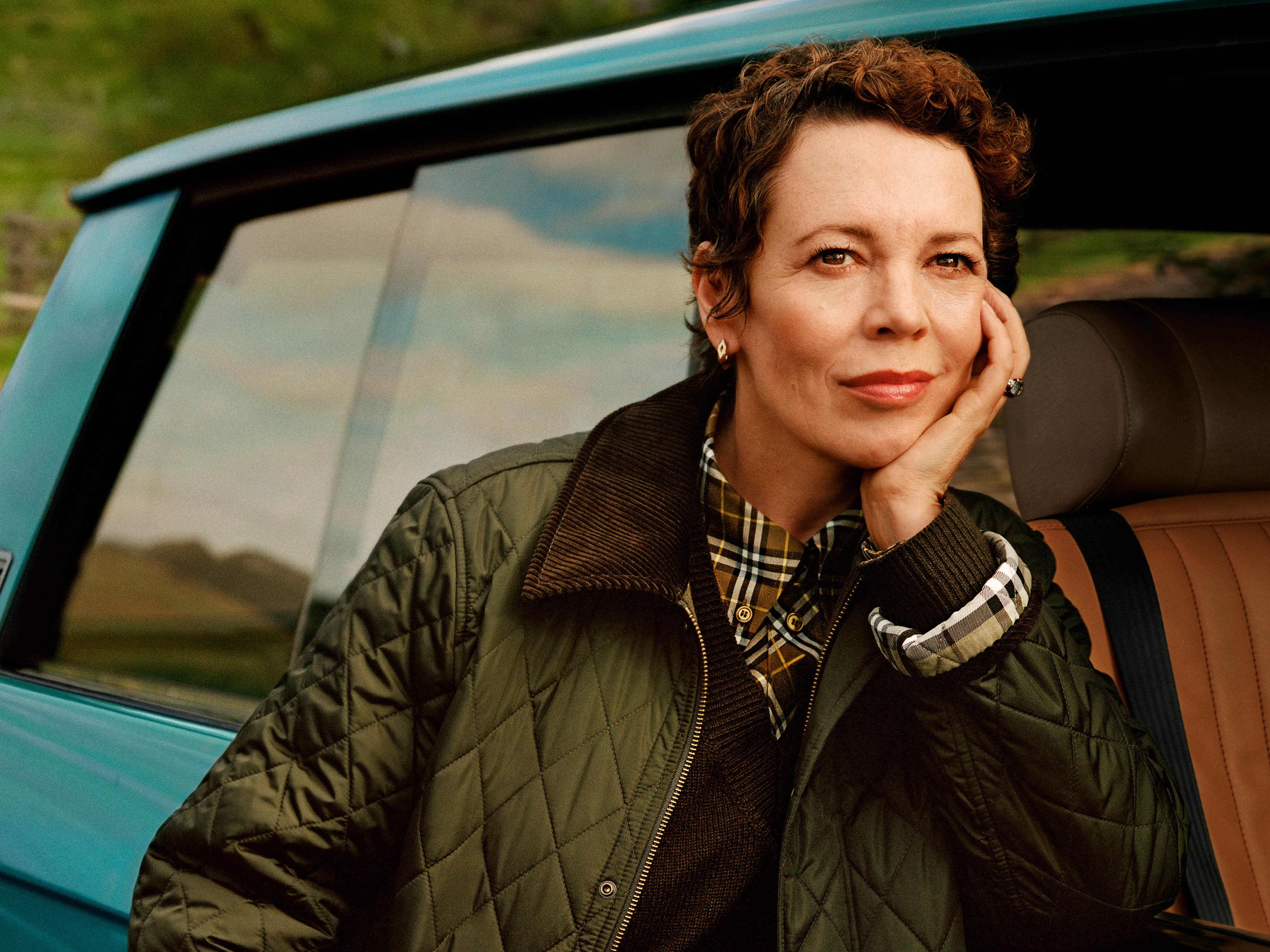 Actress Olivia Colman in a quilted Burberry jacket leans thoughtfully out of a teal car against a countryside backdrop.