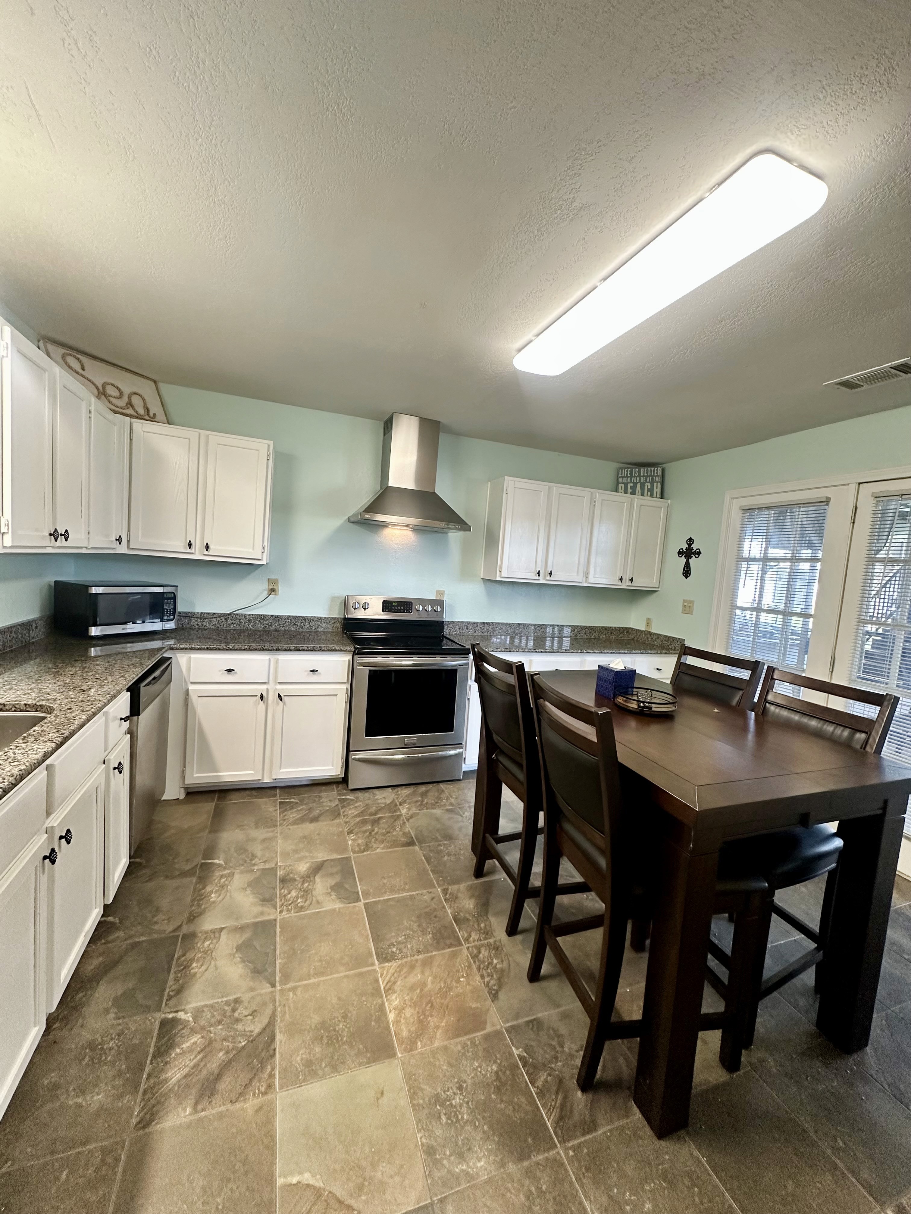 A spacious kitchen features white cabinetry, a stainless steel oven with a vent hood, granite countertops, and a large wooden dining table with high chairs against a backdrop of light turquoise walls.