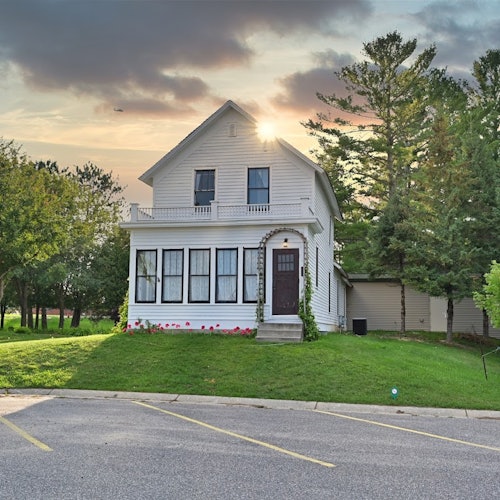 บ้านสีขาวสองชั้นพร้อมระเบียงด้านหน้า รายล้อมไปด้วยต้นไม้สีเขียวและสนามหญ้า พร้อมลานจอดรถอยู่ด้านหน้า