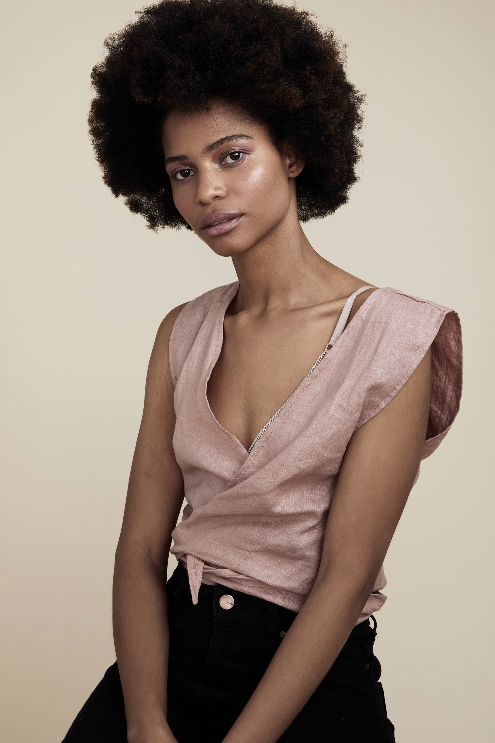 Contemporary fashion portrait of woman with afro hair wearing dusty rose linen wrap top and black high-waisted pants with zipper detail, shot against neutral background.