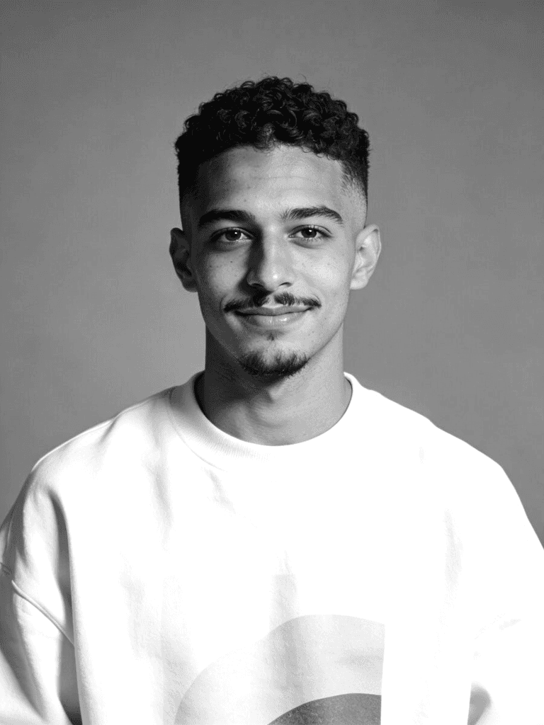Portrait of a young man with curly hair and a mustache, wearing a cream sweatshirt with a minimalist design, against a gray background.