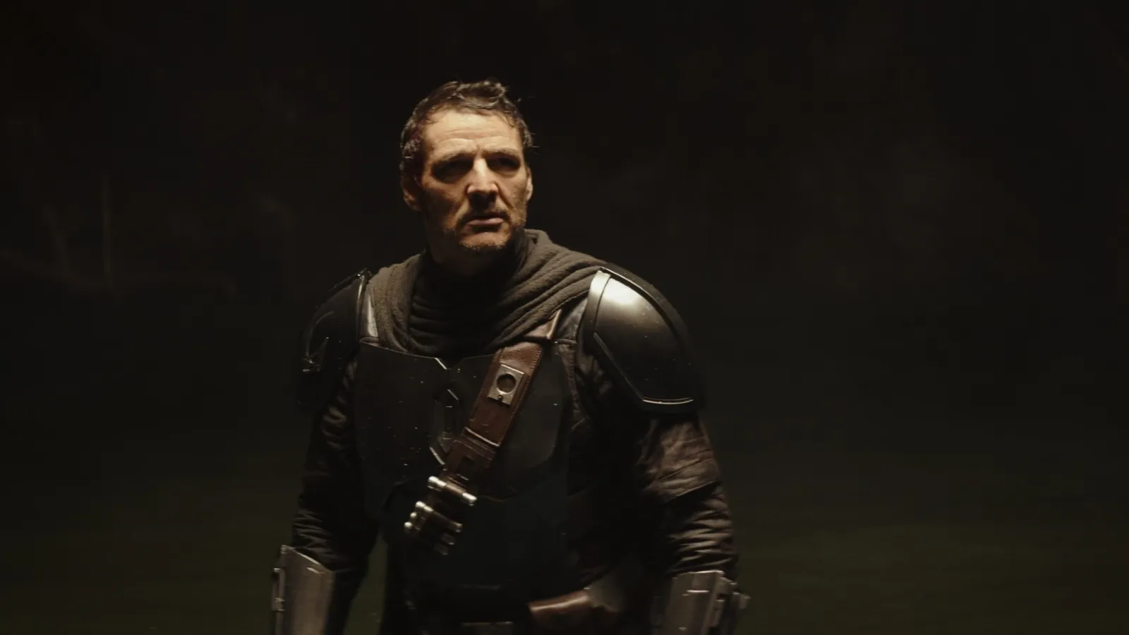 A man wearing a dark, futuristic armoured costume stands against a dimly lit background, exuding a sense of mystery and tension.