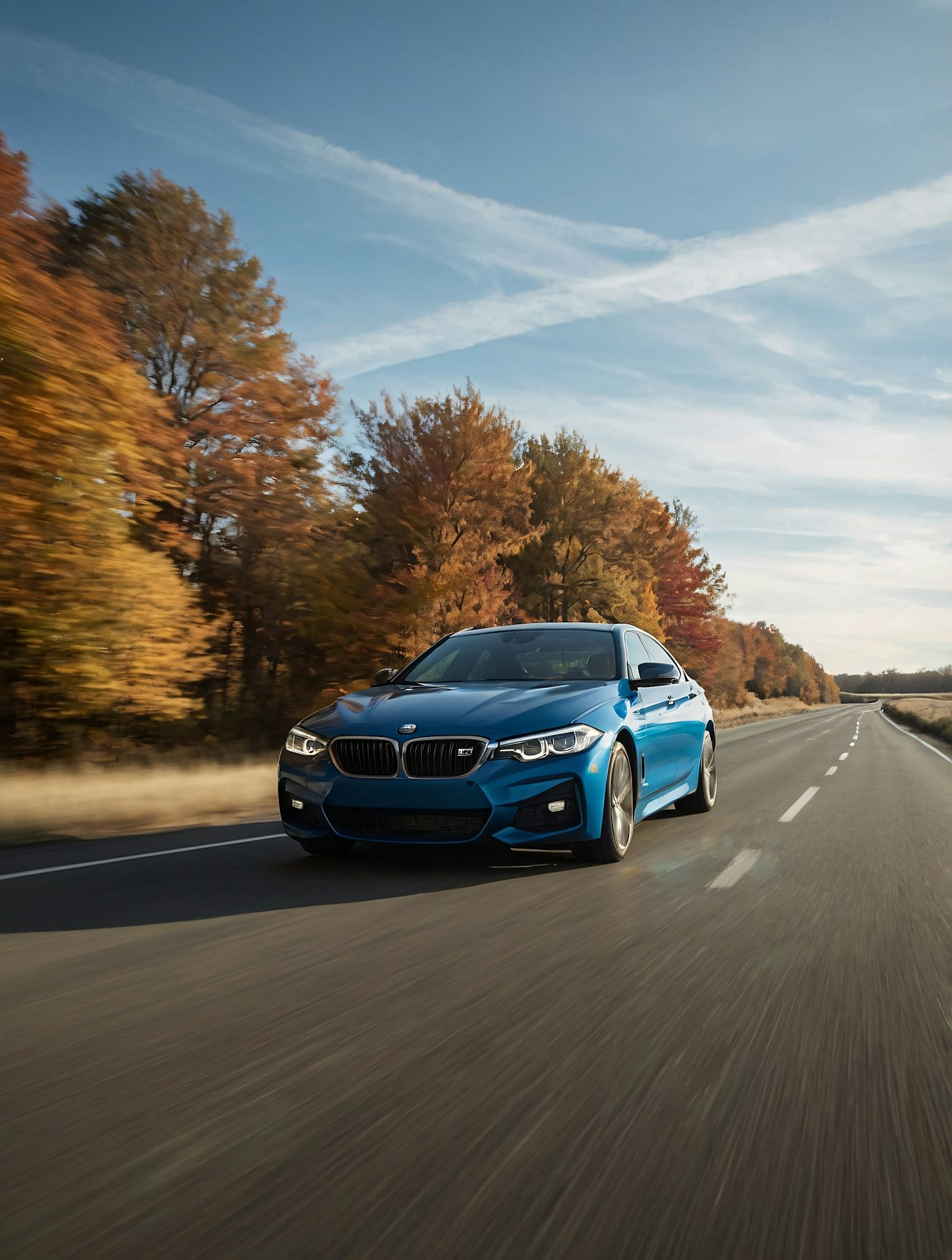 The image features a sleek, blue car driving down an open road, surrounded by a backdrop of autumnal trees with vibrant orange and yellow leaves.