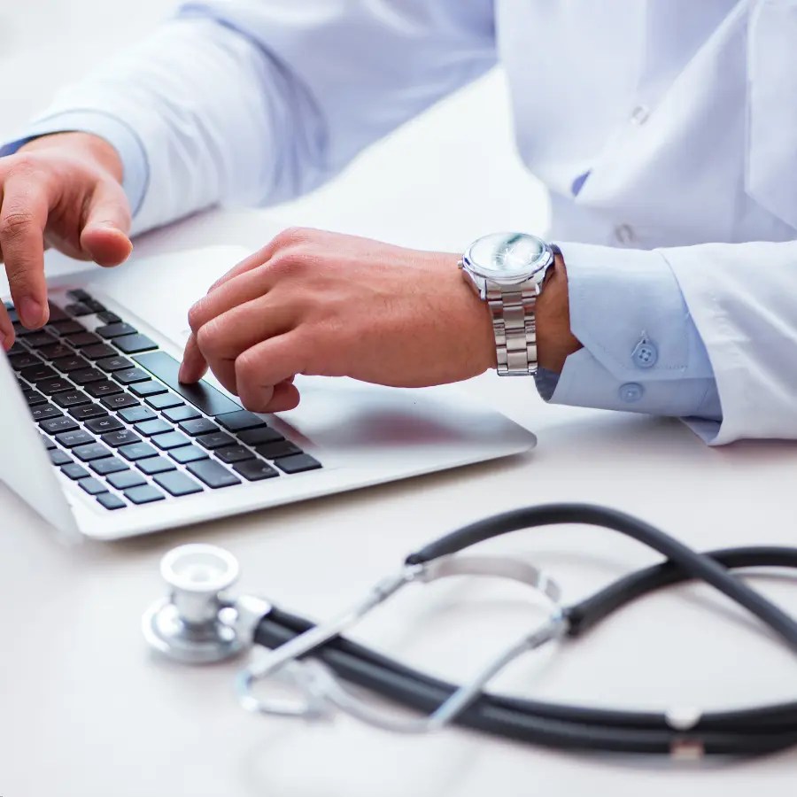 Medical dictation software assisting a doctor typing on a laptop with a stethoscope on the desk.