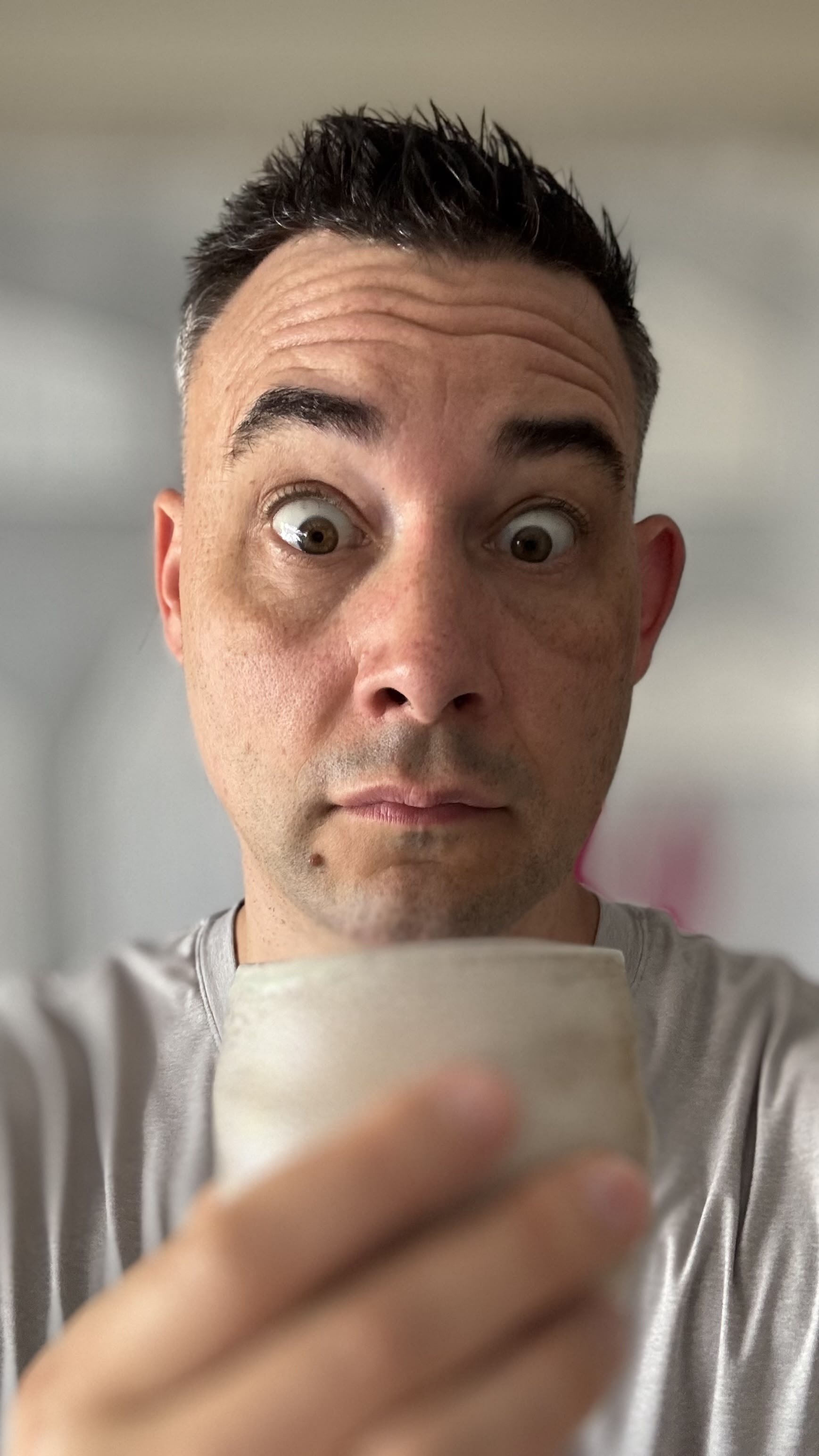 An app icon of an iPhone photo with the face of a thin man in his mid-40s, with white skin and short brown hair, holding a milk tea cup. His eyes are wide open, and his mouth is slightly opened. The background is a pink pastel color, and he has some imperfections that give him character. He's wearing light grey with a simple design, a simple color palette, simple shapes, flat colors, minimalistic, a square shape, and a circular frame border.