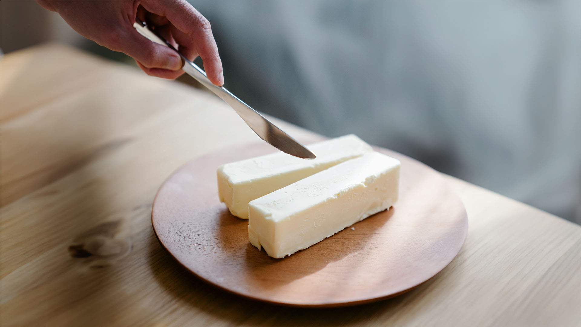 Eine Hand, die ein Buttermesser hält und sich darauf vorbereitet, ein Stück Butter zu schneiden. Die Butter liegt in zwei länglichen Blöcken auf einem runden Holzteller. Der Hintergrund ist unscharf, wodurch der Fokus auf der Hand und der Butter liegt.