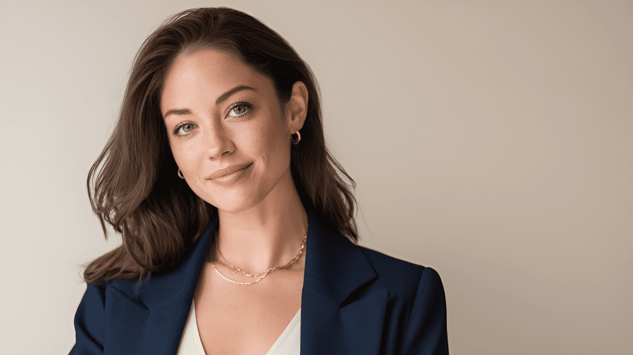 Smiling woman with long brown hair wearing a navy blazer and gold jewelry against a black background, conveying a professional and approachable tone.