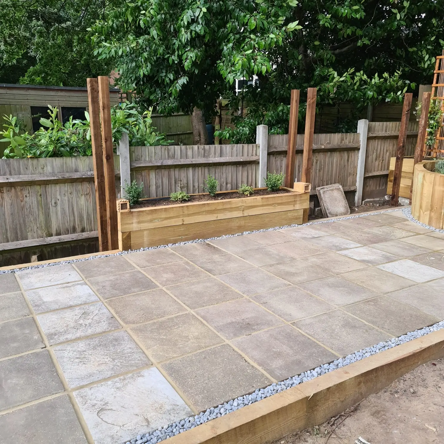 A stone patio area under construction, featuring wooden posts and a green background of trees and grass.