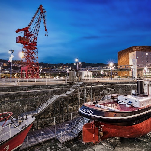 A dock scene at dusk with boats, a red crane, a bridge, and buildings in the background illuminated by streetlights.