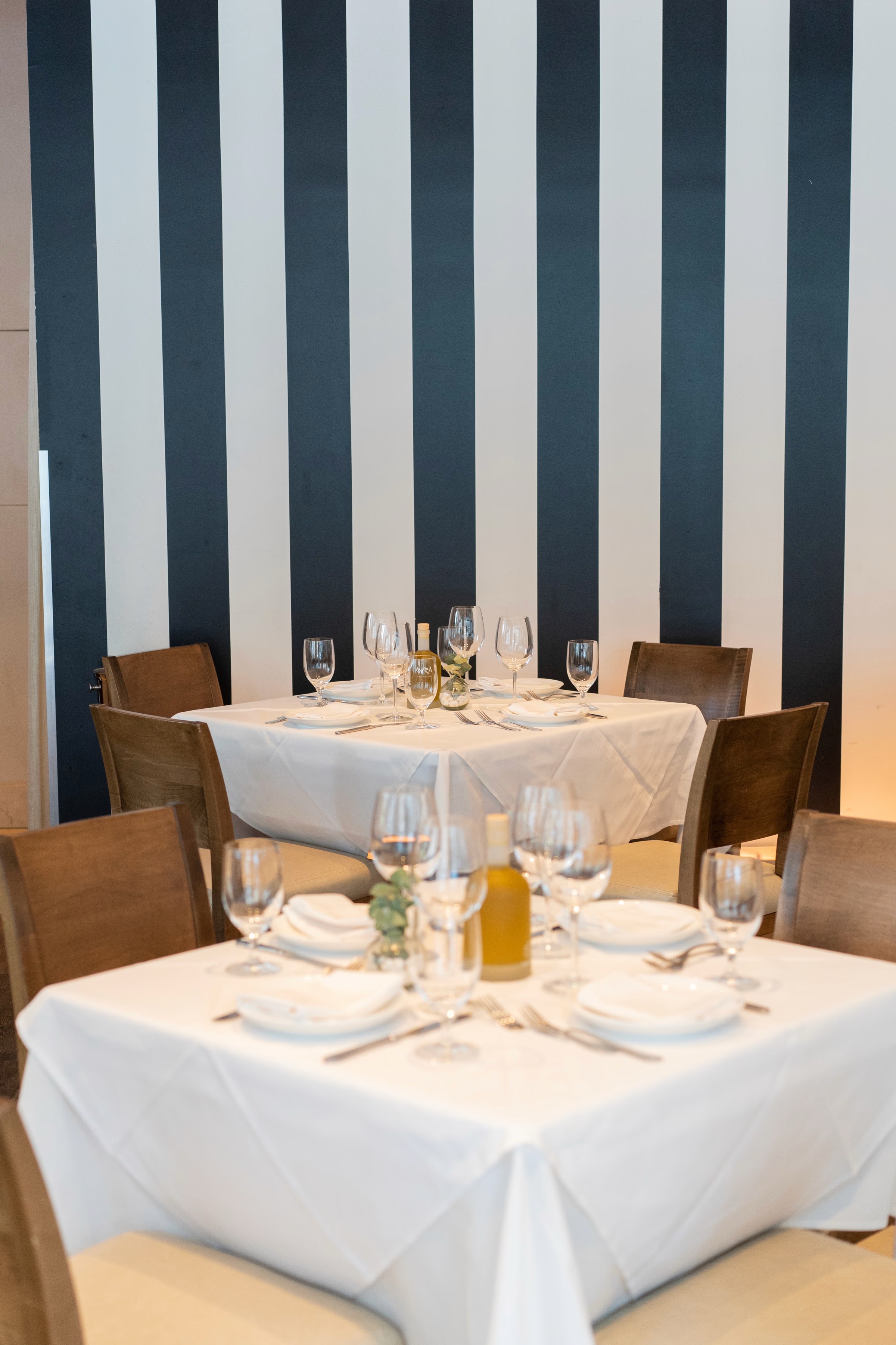A neatly set dining table with white tablecloth, surrounded by chairs, against a backdrop of vertical stripes.