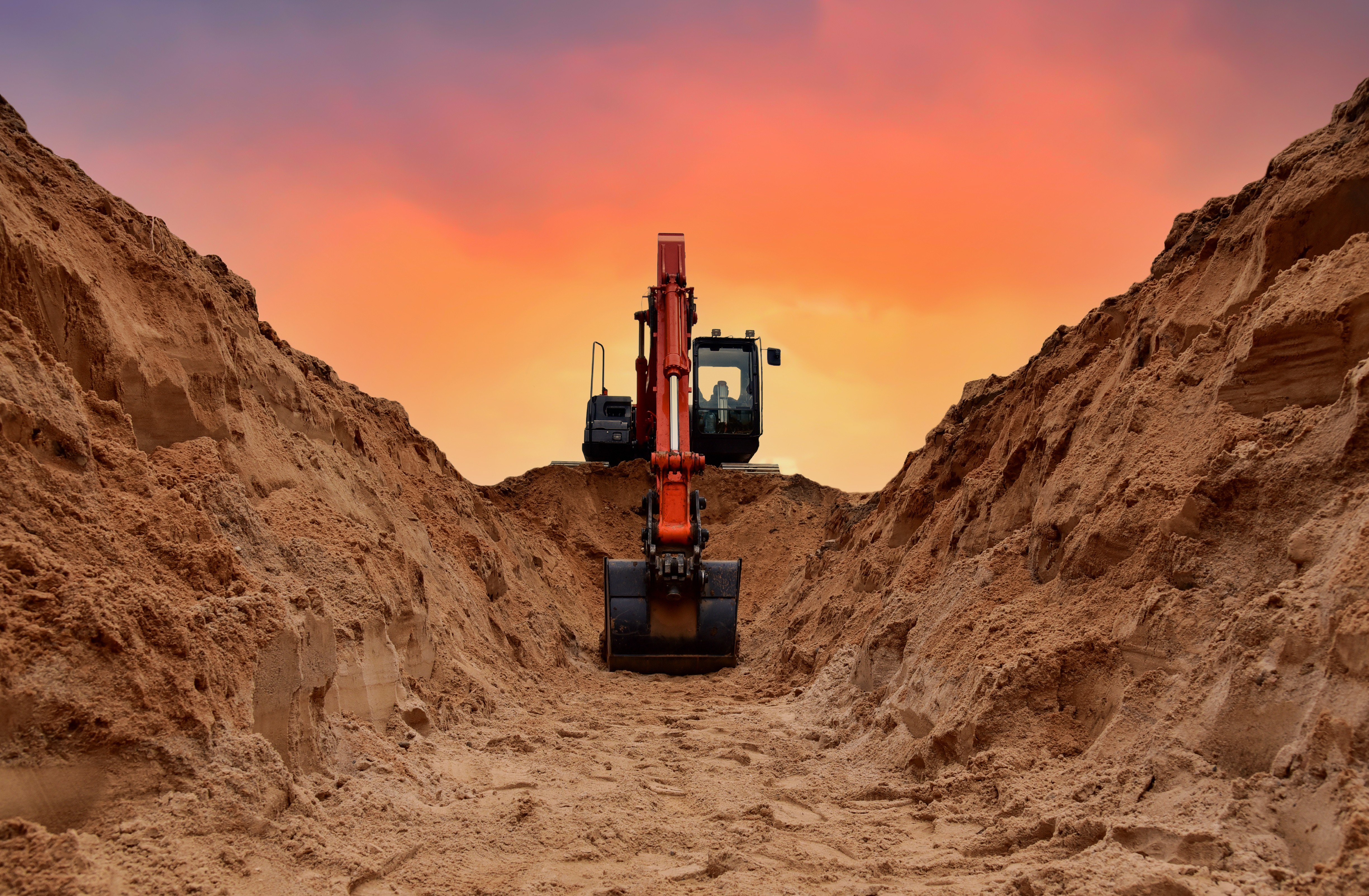 Front view of an excavator digging between tall mounds of soil at a mining site during a vibrant sunset.