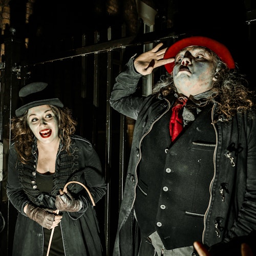 연극 분장과 의상을 입은 두 사람이 어두운 막대 배경 앞에서 나란히 서 있으며, 그중 한 명은 빨간 모자를 쓰고 있습니다.