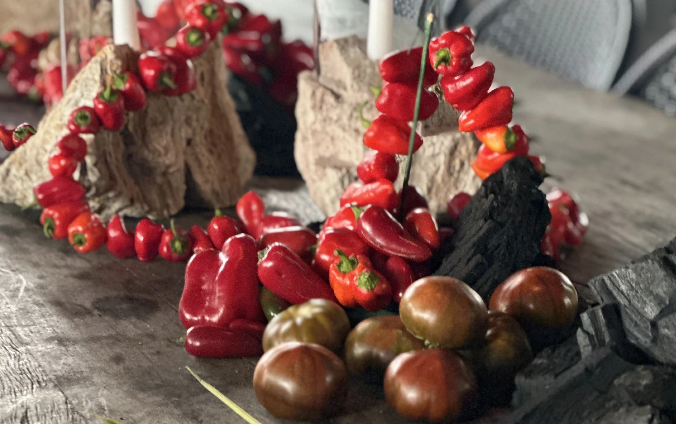 Puesta de mesa con ajíes picantes, tomates heirloom oscuros, carbón y velas, celebrando la materia prima.