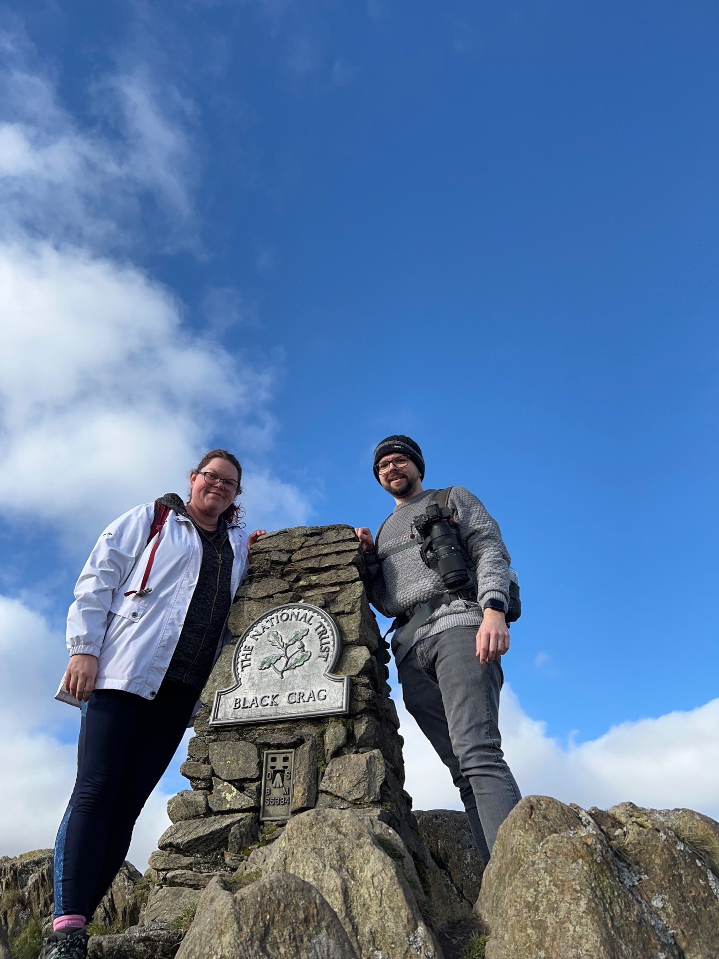 A low, wide-angle shot of Martin & April stood next to the Black Fell cairn (the plaque actually reads Black Crag)