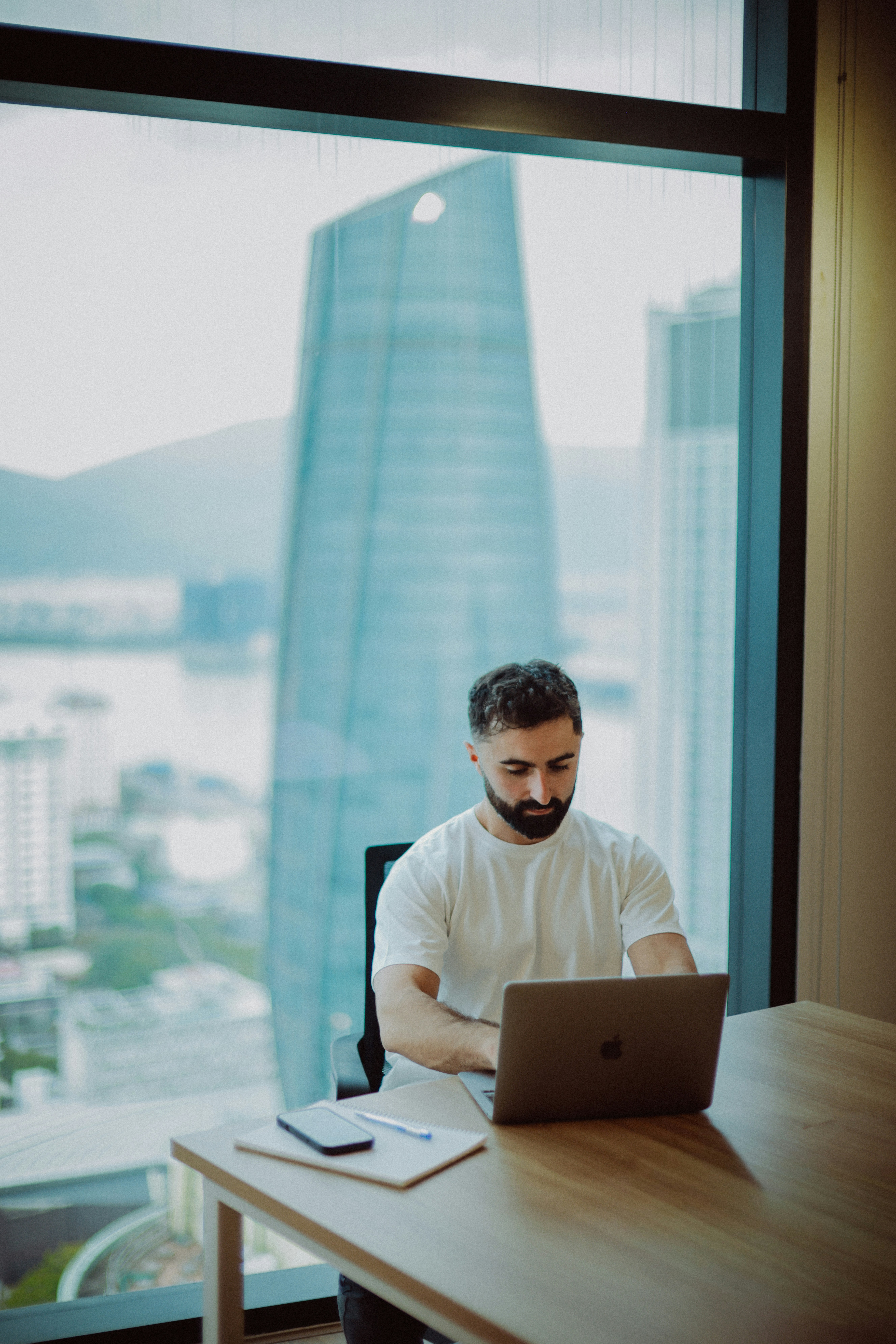 Man working on a laptop by a large window.